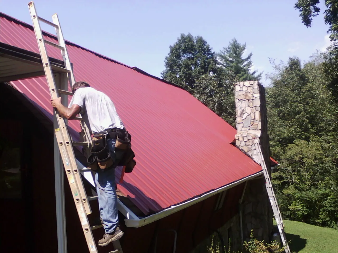 Roofer on a ladder beside a red metal roof and stone chimney — Soffit & Fascia Repair by Roofing Brothers in Aston